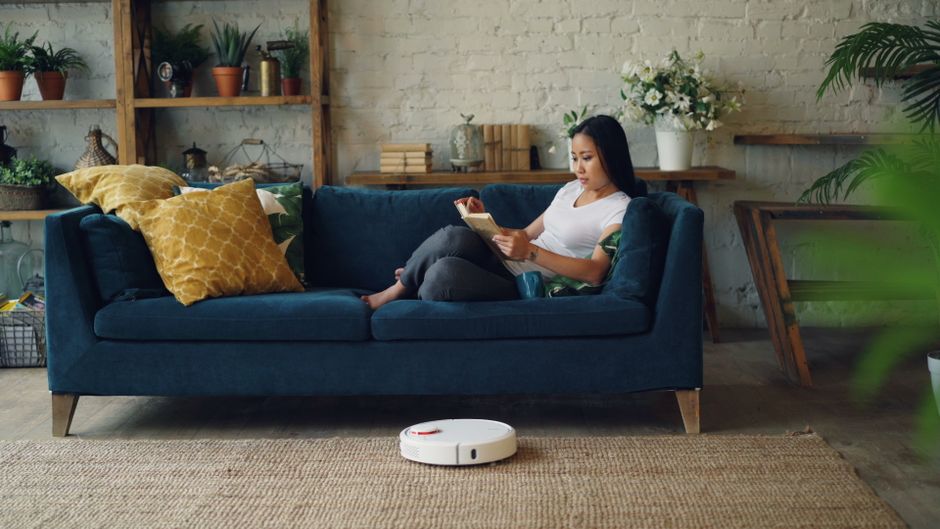 A woman sitting on a deep blue fabric sofa with wooden legs in a well-lit living room, using a tablet. The sofa is decorated with mustard yellow cushions, and a white robot vacuum cleaner is on a beig
