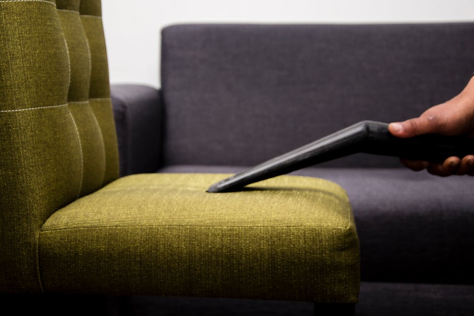 A woman sitting on a deep blue fabric sofa with wooden legs in a well-lit living room, using a tablet. The sofa is decorated with mustard yellow cushions, and a white robot vacuum cleaner is on a beig