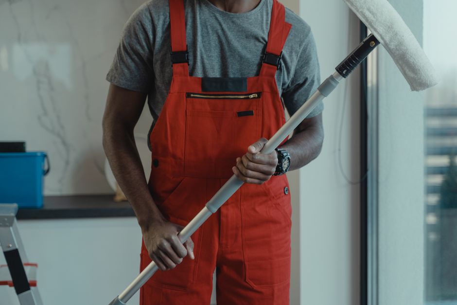A professional cleaner from Parsons Green Carpet Cleaners is holding a mop with a white head and black handle, positioned upright in a modern, well-lit interior. The person is wearing a grey t-shirt a