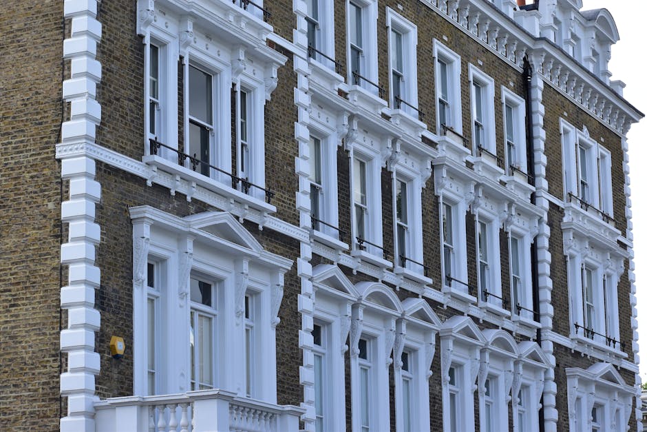 Photograph of a multi-story residential building with a classic façade, featuring brick walls and ornate white window framing. The building has several large windows with white wooden frames and decor
