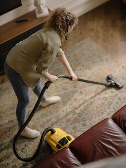 A man with curly dark hair and a beard is bending down on a wooden floor in a well-lit room, adjusting or preparing a beige woven carpet or rug on the floor. He is dressed in a bright orange long-slee